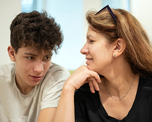 A young man and his parent sit next to each other in a classroom and talk
