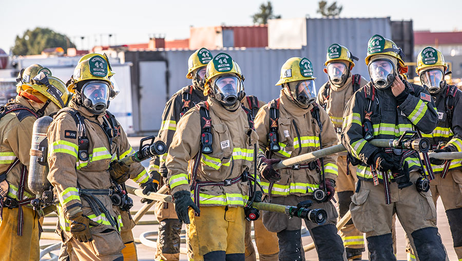 A group of firefighter cadets in full protective gear on the training grounds, holding fire hoses