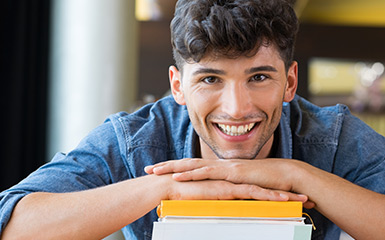 A smiling student rests their head and hands on a small pile of books